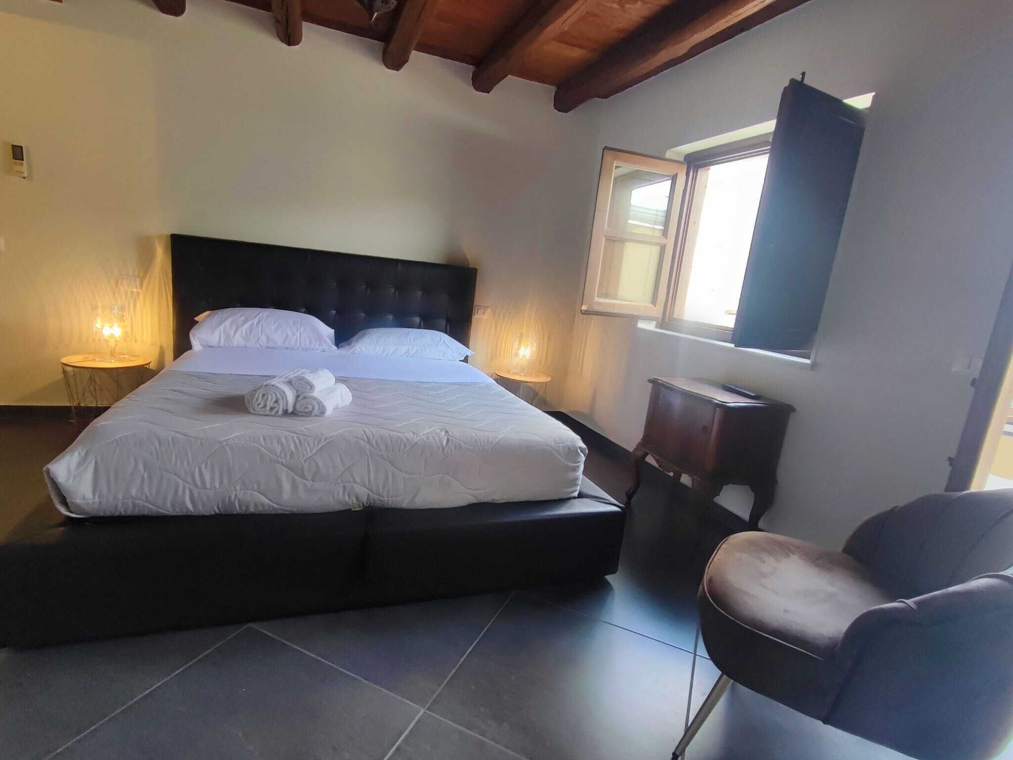 Una cama bien tendida con sábanas blancas y toallas enrolladas, un cabecero oscuro y una mesita de noche con una lámpara.