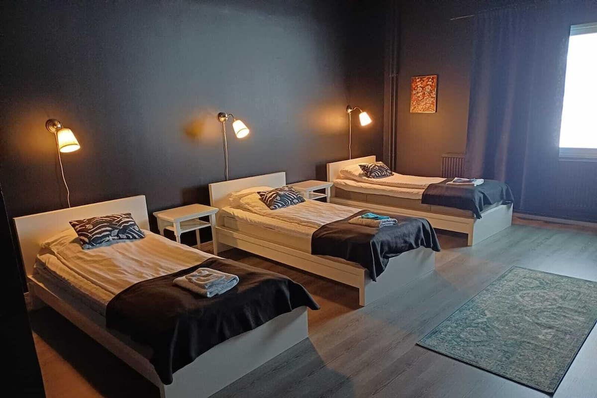 Una habitación de hotel moderna con tres camas individuales, cada una con una mesita de noche y lámpara. La habitación tiene una pared oscura y un ventanal grande a la derecha.