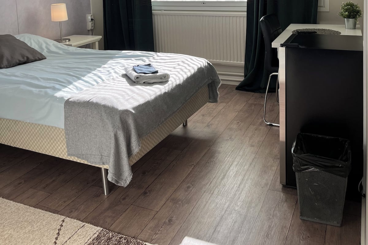 Habitación de hotel con cama, escritorio, silla, ventana con cortinas, radiador y luz en el techo.