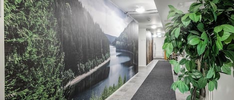Un pasillo con un mural que muestra un bosque y un río, una planta en maceta y suelo cubierto de alfombra.