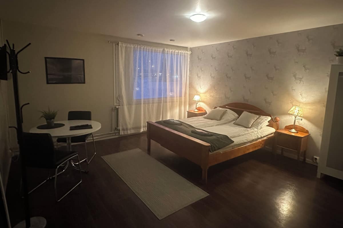 Un dormitorio con cama, mesitas de noche con lámparas, una silla, una pequeña mesa y una ventana con cortinas.