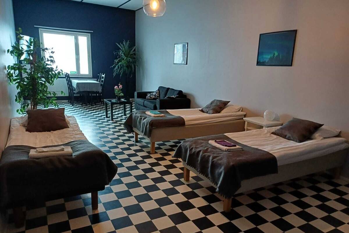 Habitación con piso a cuadros, dos camas, un sofá, una mesita y una silla. Se aprecia una ventana con vista a árboles y una planta en la esquina.