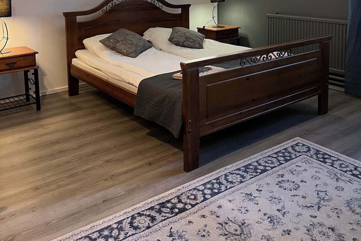 Un dormitorio con cama de madera, mesita de noche, lámpara y alfombra estampada.
