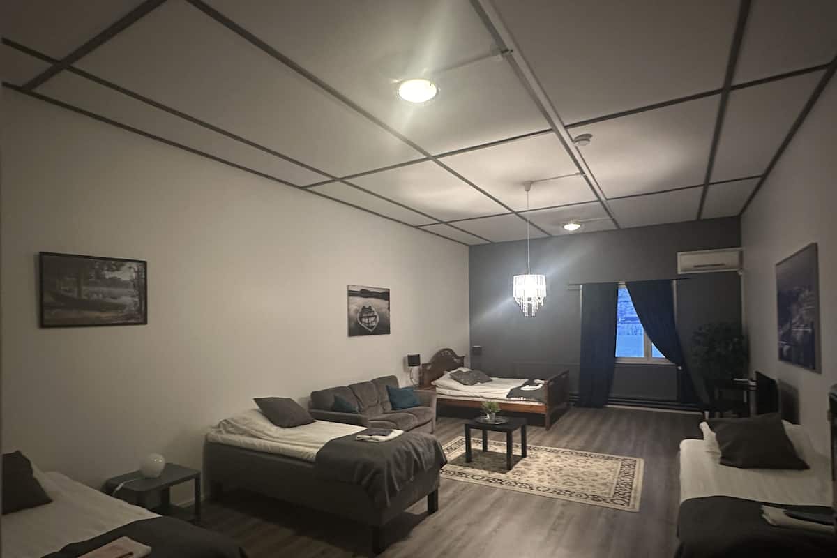 Una habitación moderna y luminosa con cama, sofá y una pequeña mesa. Se observa una araña, una ventana con cortinas y obras de arte en las paredes.