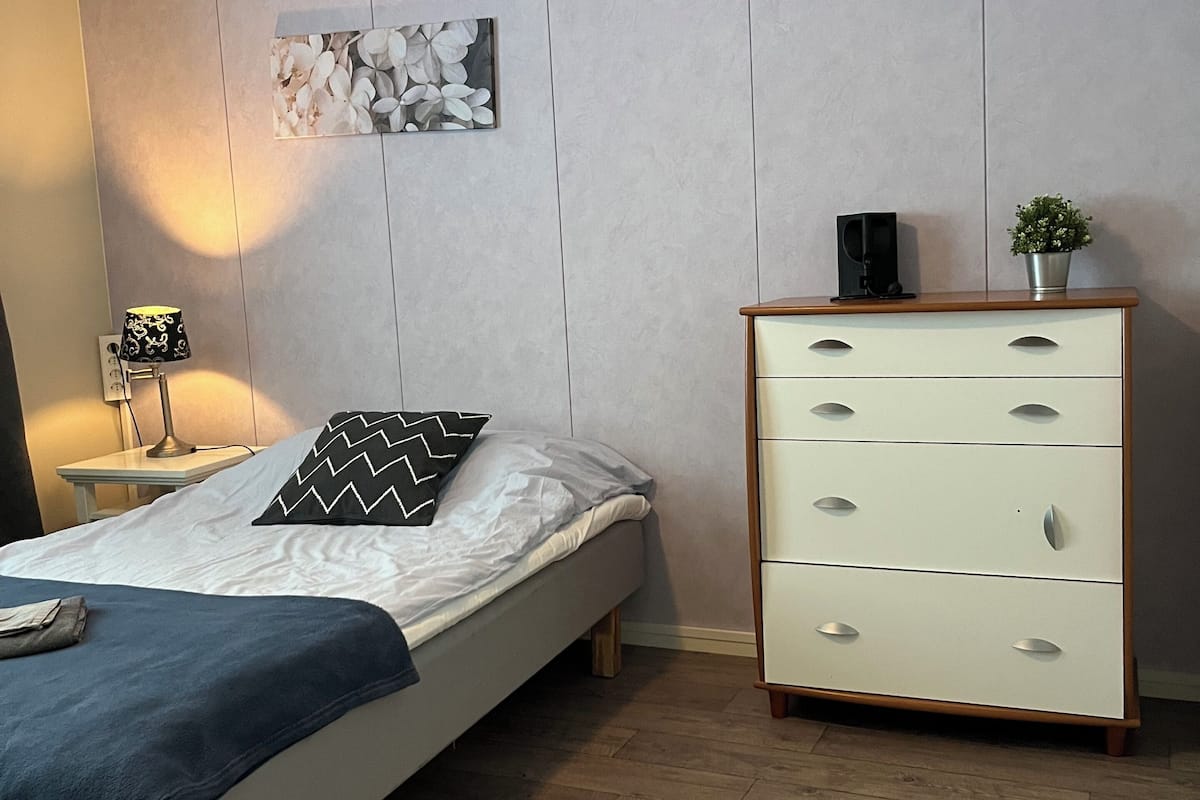 Un dormitorio con cama, mesita de noche, lámpara, cómoda y un cuadro colgado en la pared.