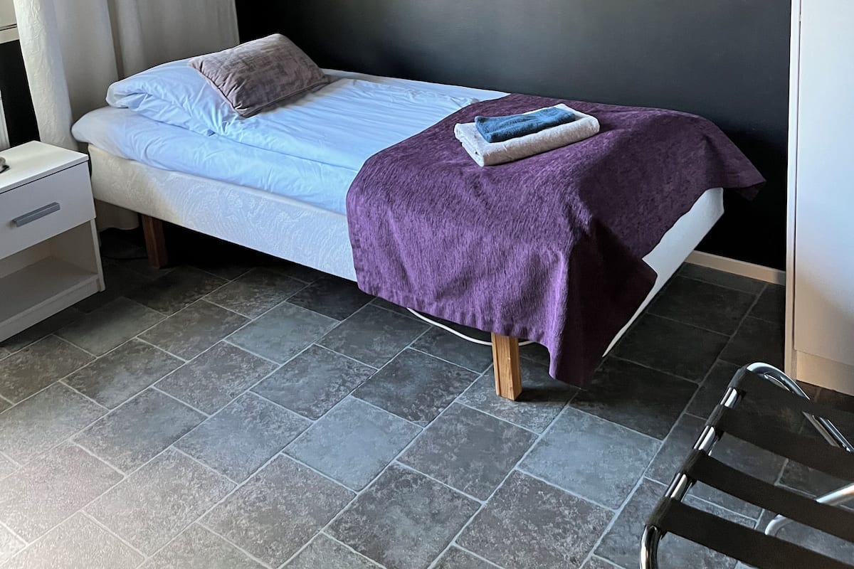 Una cama individual con cabecera blanca, una colcha morada y un almohadón. Se observa un mesita de noche blanca con un cajón y una lámpara. La habitación tiene paredes oscuras y piso de baldosas.