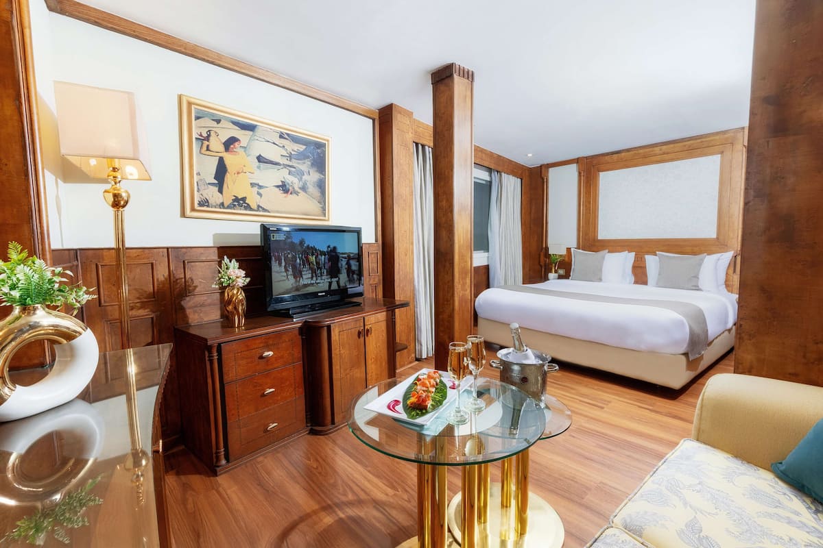 A hotel room with a bed, a TV, a glass table with a plate of food and drinks, wooden furniture, and a painting on the wall.
