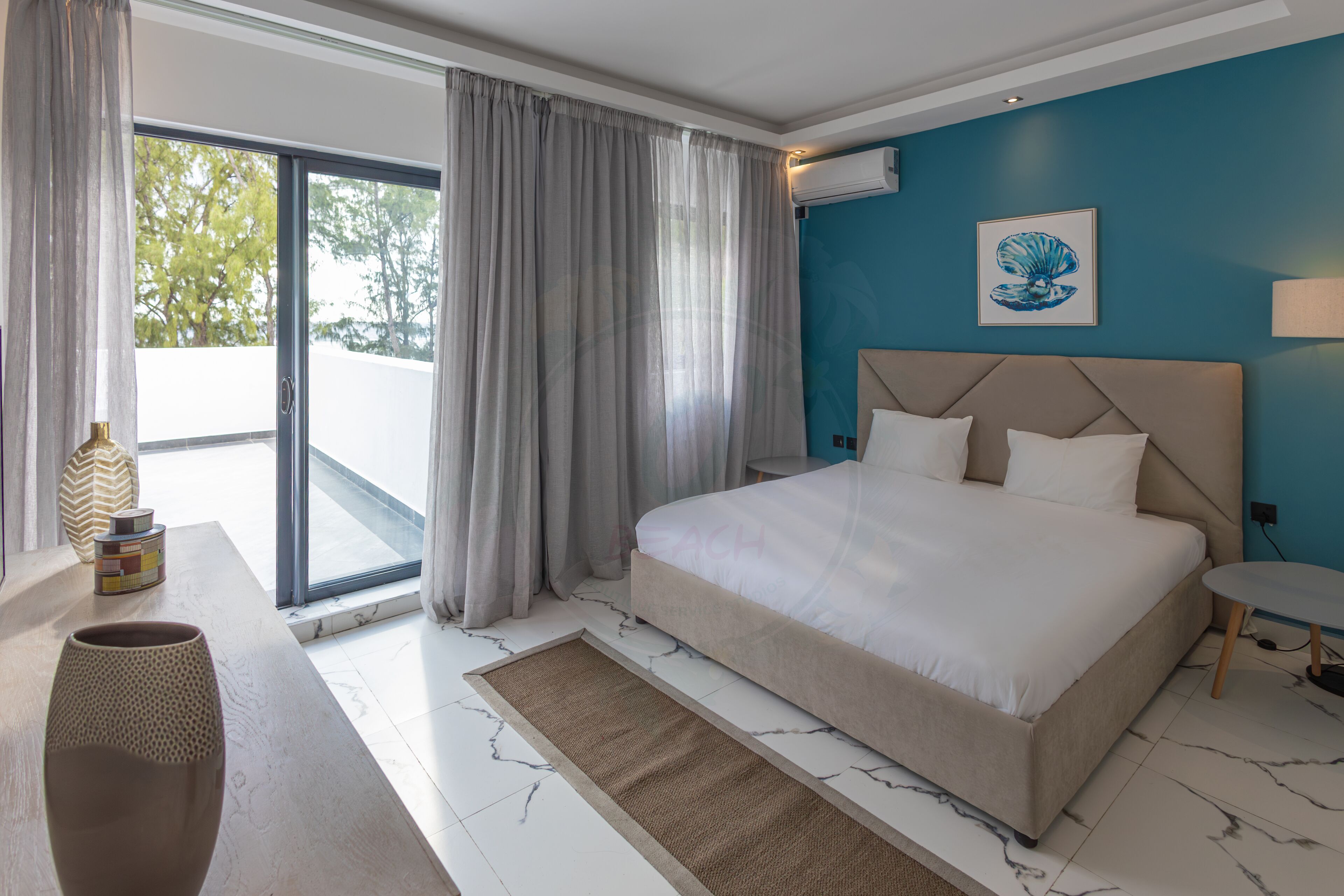 A modern hotel room with a large bed, a view through a sliding glass door, a blue accent wall, and a decorative vase on the nightstand.