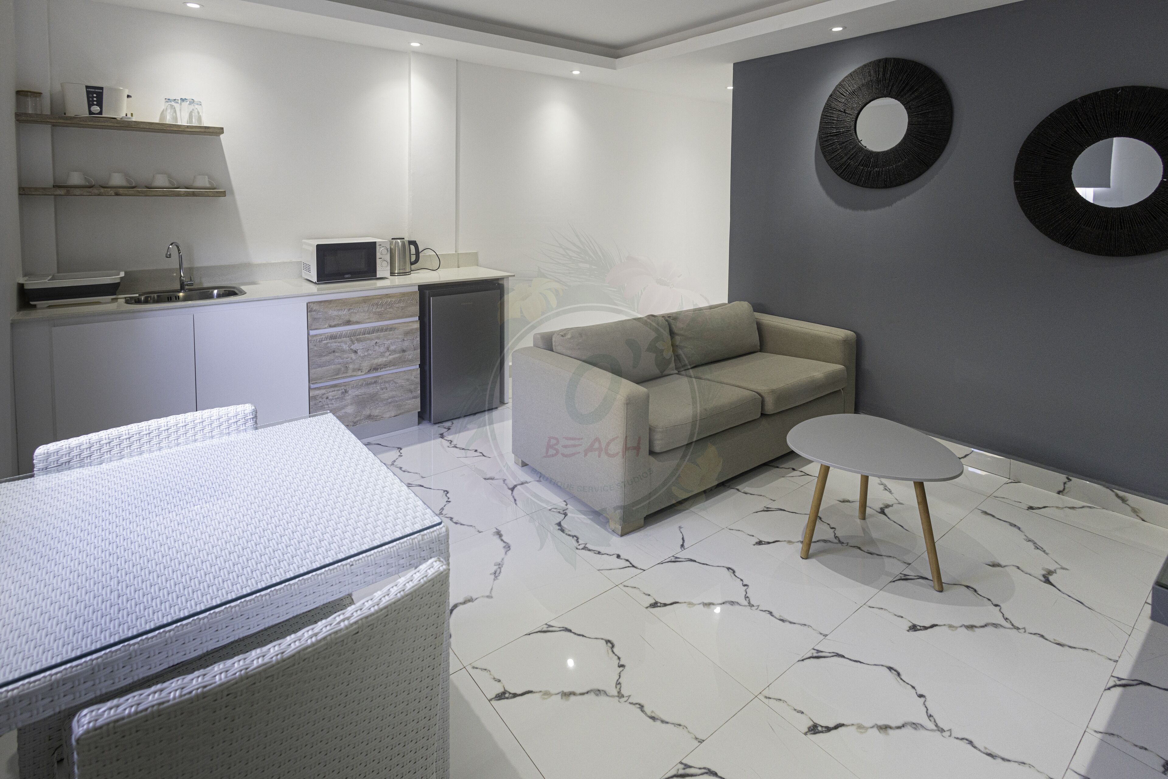 A modern kitchen with white cabinets, a microwave, and a sink. There is a beige sofa and a small round table in the living area.
