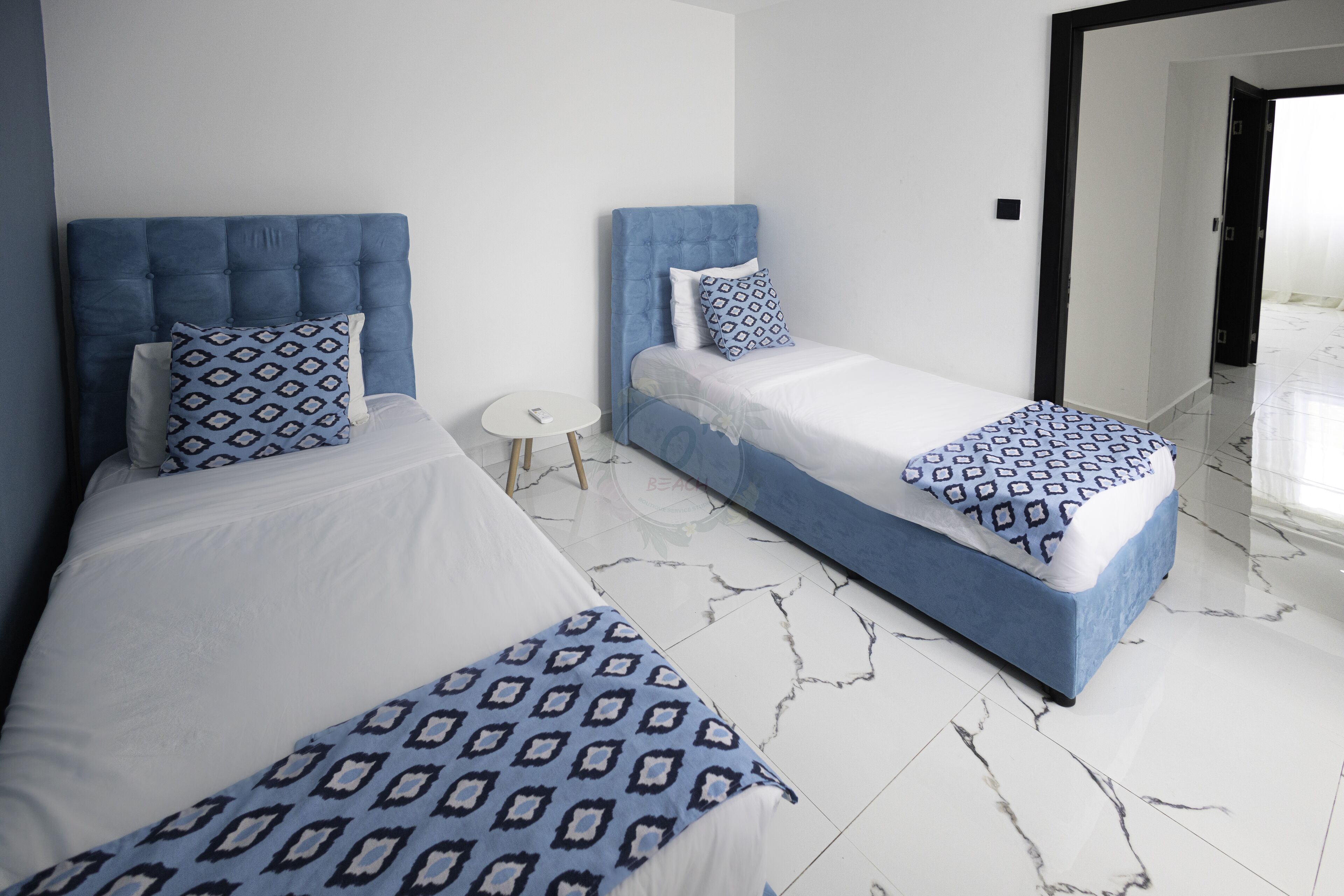 A hotel room with two beds, each with a blue headboard and patterned pillows. The room has white walls and a marble floor.