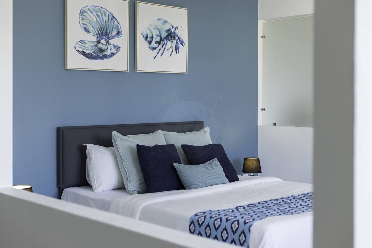 Ein ordentlich bezogenes Bett mit einer gemusterten Decke, einem dunklen Kopfteil und mehreren Kissen in Blau- und Weißtönen. Darüber befinden sich zwei gerahmte Kunstwerke, und eine Nachttischlampe ist sichtbar.
