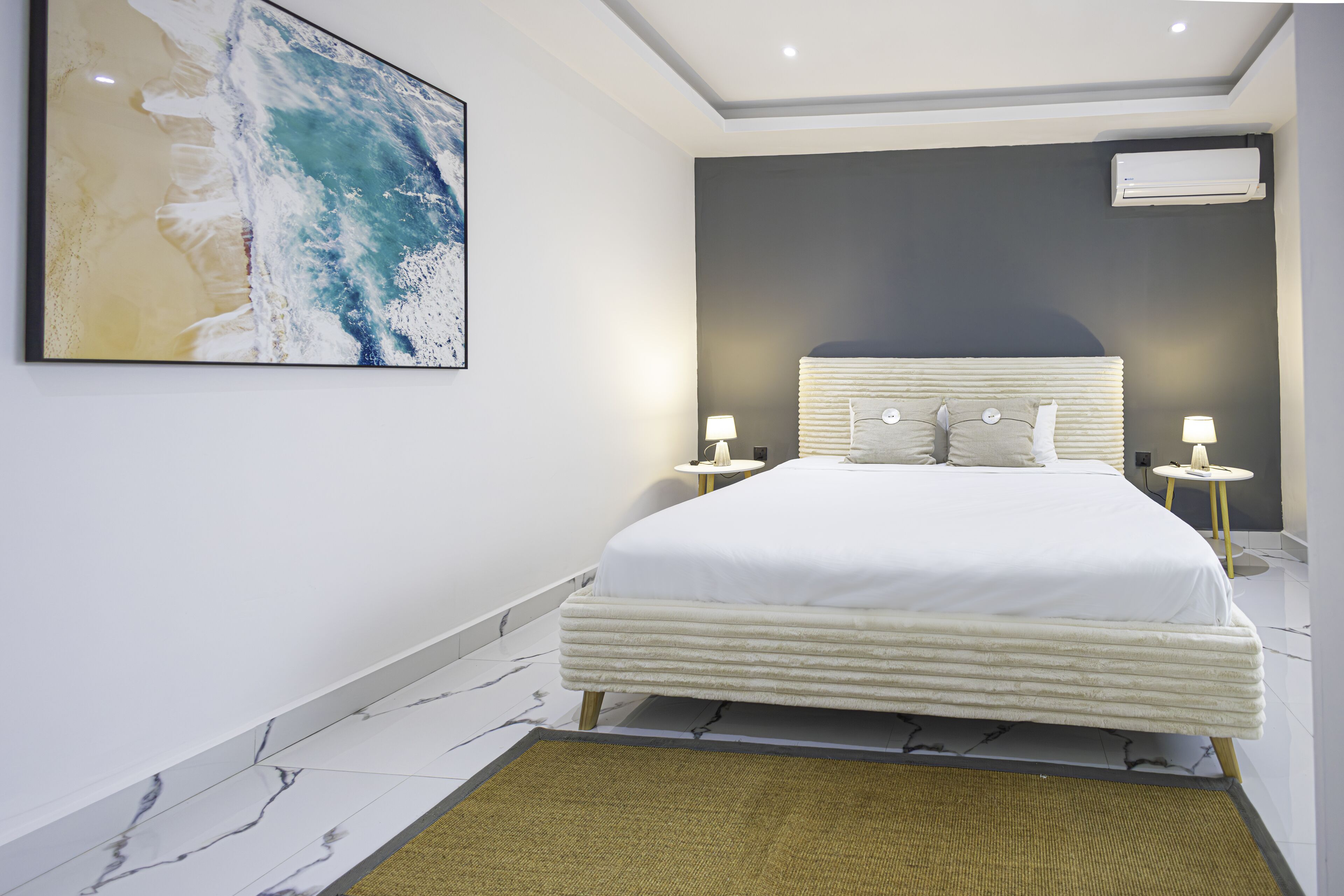 A modern bedroom with a large bed, marble flooring, a beach-themed painting, and an air conditioning unit.