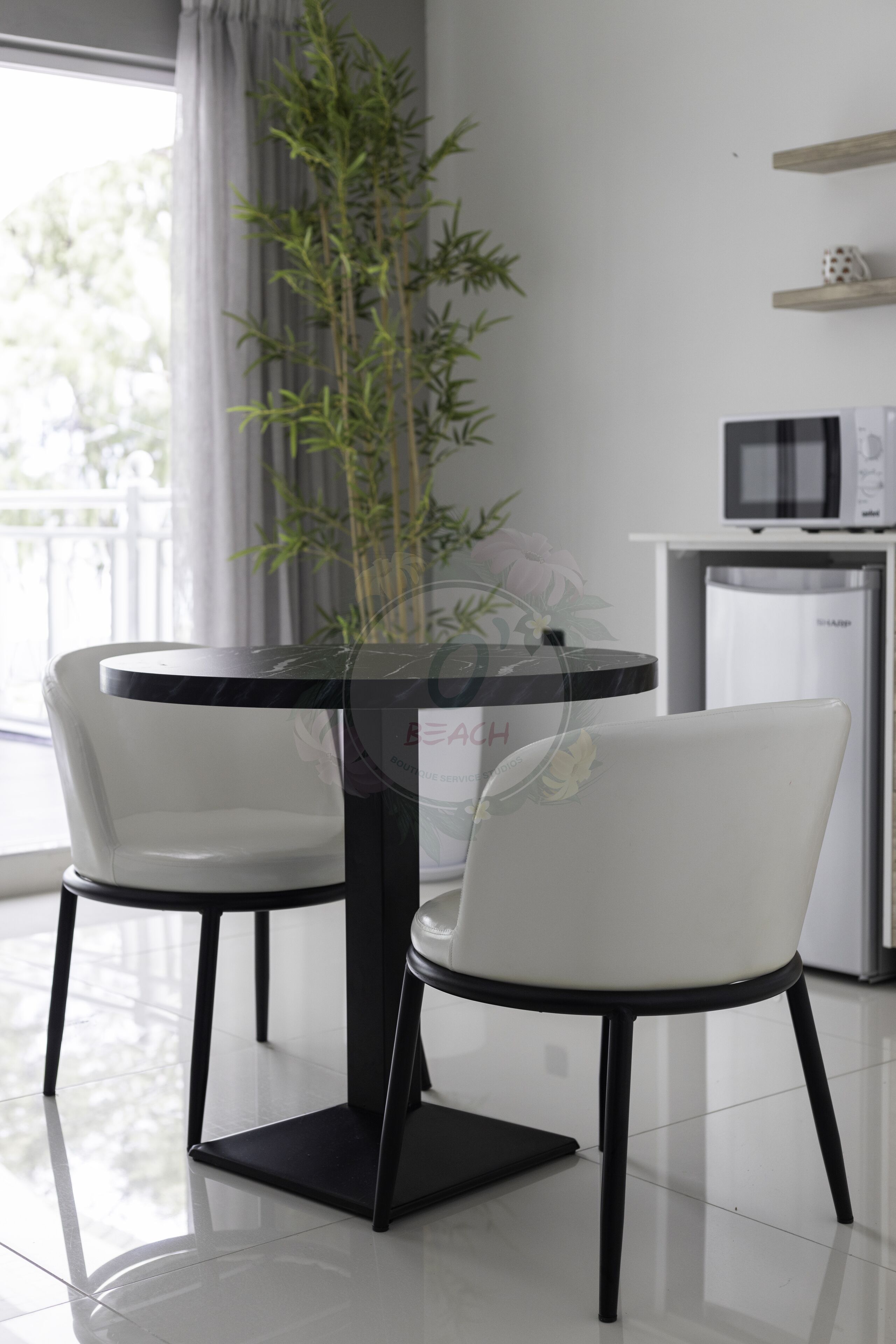 Et moderne spiseområde med et rundt bord og to hvide stole, en glasvase med en plante, og et køkken i baggrunden.
