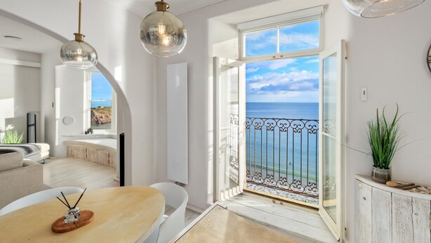 A modern living room with a large window offering a view of the sea, a dining area with a table and chairs, and a kitchen visible in the background. - Min Y Mor - Breathtaking Sea Views (Tenby)