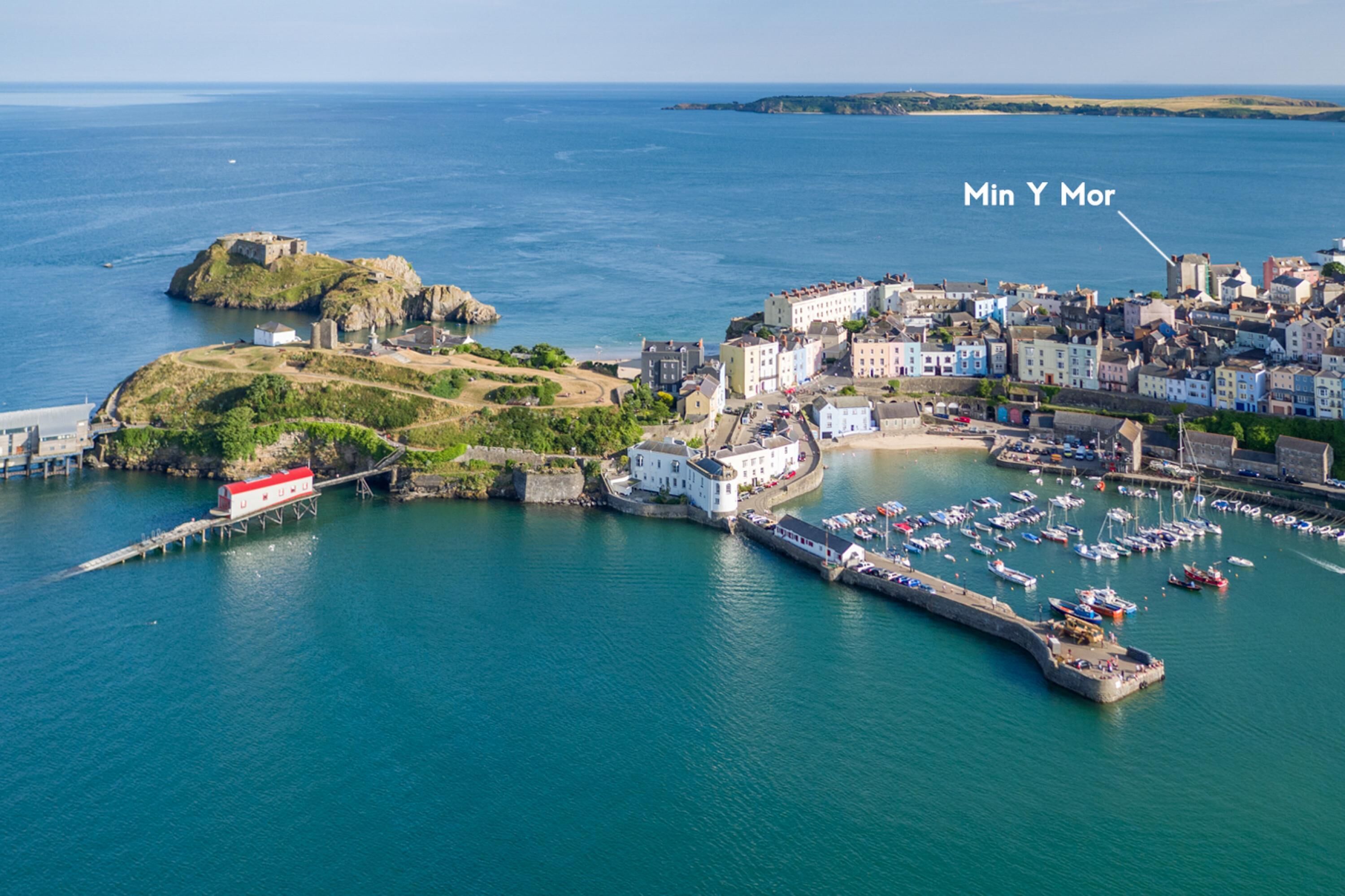 An aerial view of a coastal town with a prominent castle, a marina with numerous boats, and a mix of historic and modern buildings.