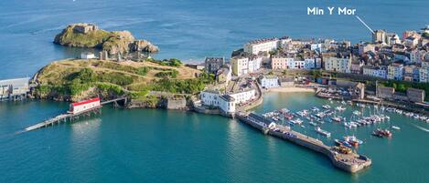An aerial view of a coastal town with a prominent castle, a marina with numerous boats, and a mix of historic and modern buildings.
