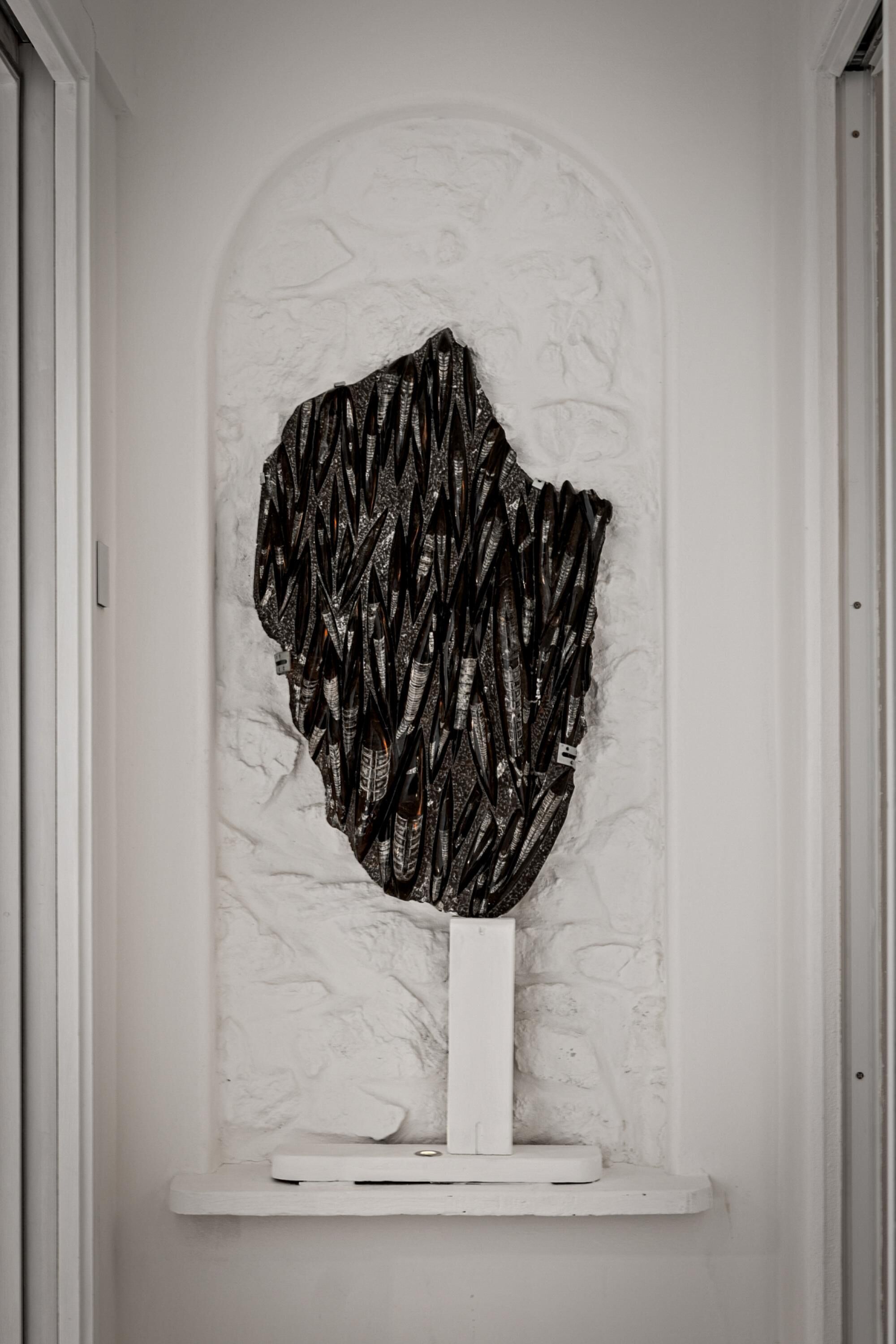 A white wall with a textured pattern and a black abstract sculpture mounted on a white pedestal.
