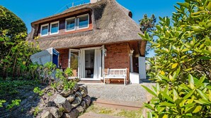 Une maison à toit de chaume avec un rez-de-chaussée en briques, un banc en bois à l’extérieur et un mur de pierre au premier plan.