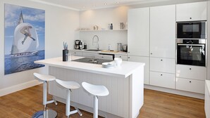 Une cuisine moderne avec des meubles blancs, un îlot central doté de tabourets, une grande peinture représentant un voilier et des appareils électroménagers intégrés.