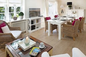 Ein helles, offenes Wohnzimmer mit Esstisch, Korbmöbeln, einem Fernseher und einem Bücherregal.