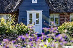 Uma casa com fachada azul, porta e janelas brancas, cercada por um muro de pedra e plantas floridas.