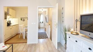 Una cocina moderna con armarios blancos, un comedor con mesa y sillas blancas, y una sala de estar con televisor y objetos decorativos.