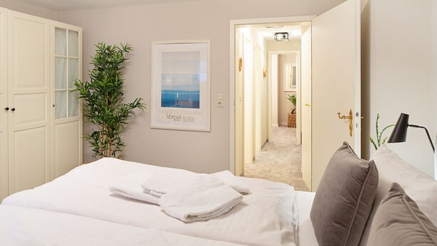 Una habitación de hotel con cama, toallas blancas, un cartel, una planta y una puerta que da a otra habitación.