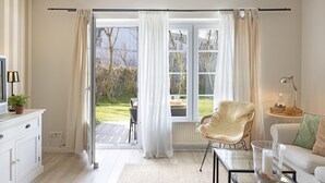 Una sala moderna con una mesa de centro de vidrio, un sofá blanco, un sillón de mimbre y vistas a un jardín a través de amplios ventanales.