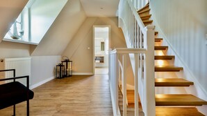 A spacious attic room with a sloped ceiling, wooden flooring, a white staircase with a railing, and a view through an open door to another room.