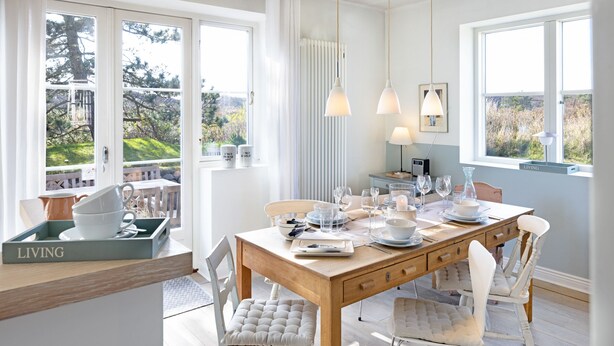 A dining area with a wooden table set for four, white upholstered chairs, and a view of the outdoors through large windows.