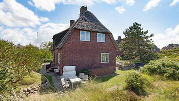 Een huis van rode bakstenen met een steile puntige kap, een klein portaaltje met een bankje en een tuin met buitenmeubilair.
