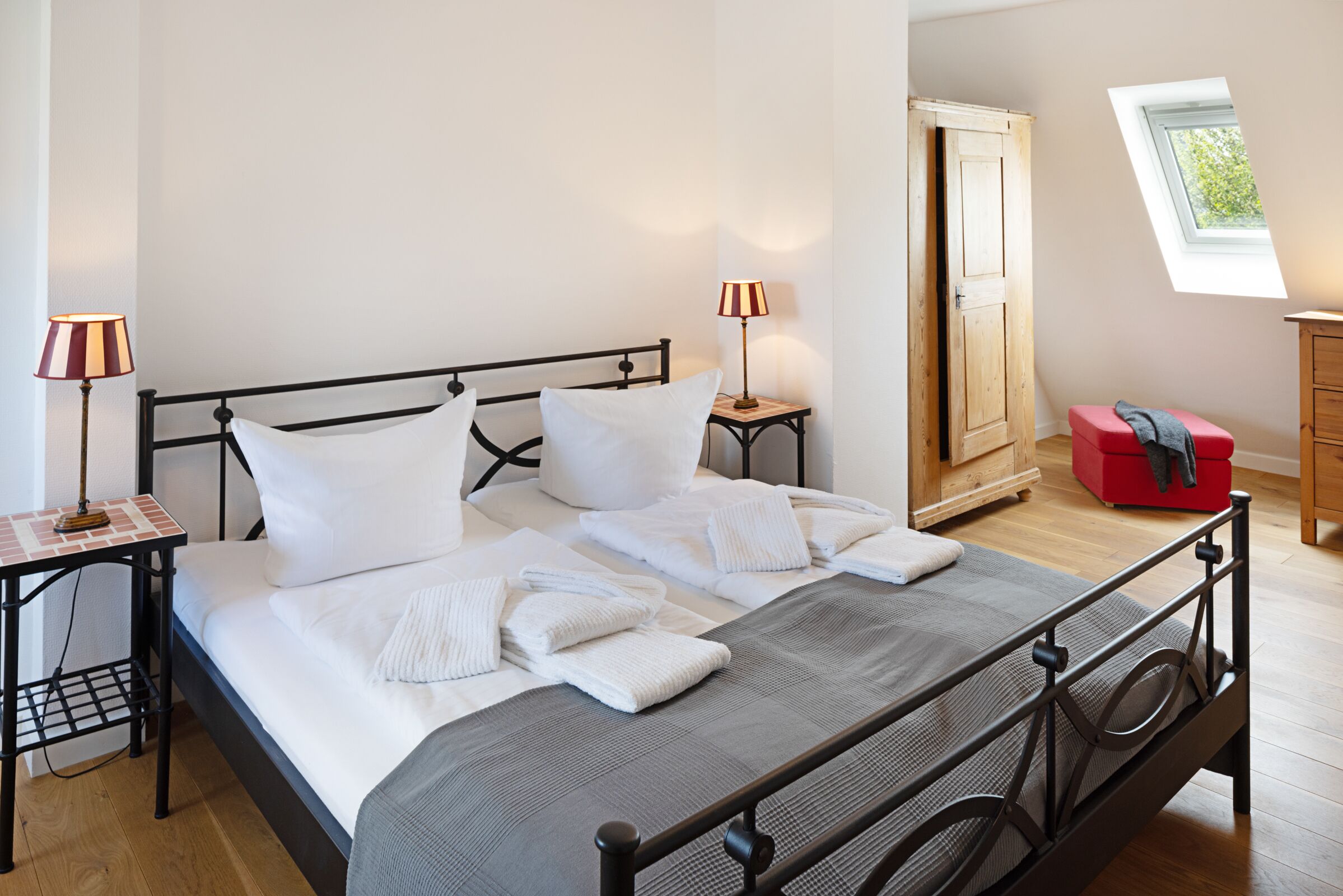 Een net opgemaakt bed met witte lakens, een nachtkastje met een lamp, en een raam met uitzicht op groen.