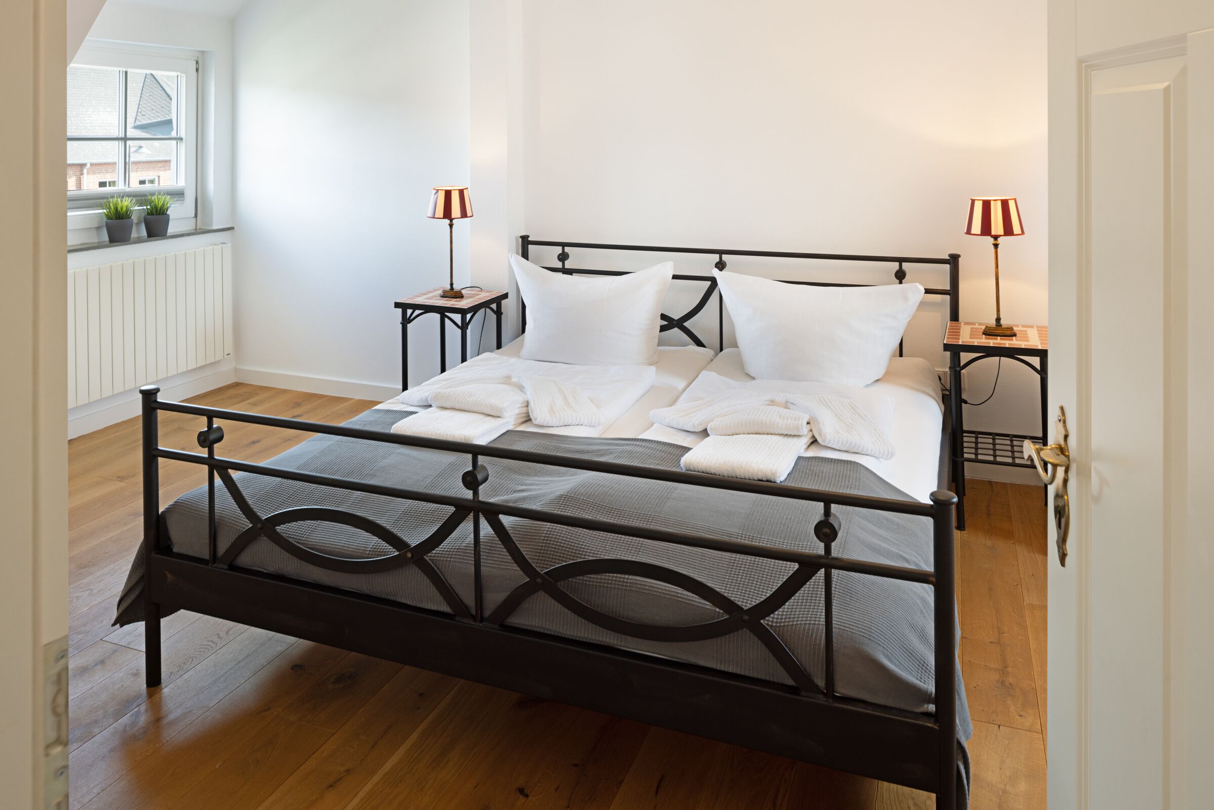 Una cama bien hecha con sábanas blancas, dos mesitas de noche con lámparas y una ventana con plantas.