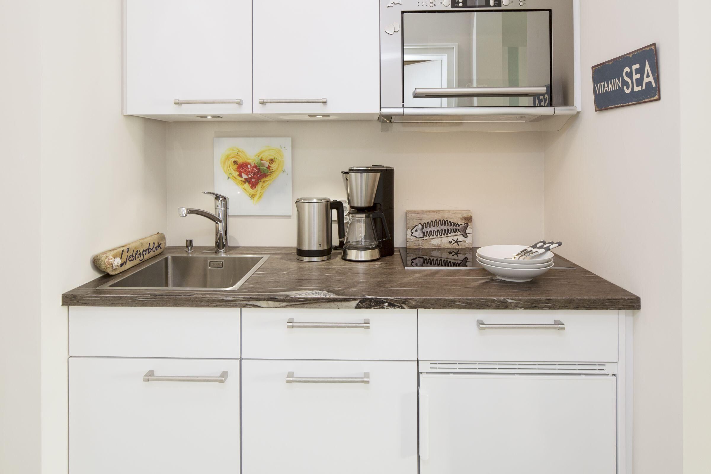 A kitchen with white cabinets, a stainless steel sink, a coffee maker, and a heart-shaped artwork on the wall.