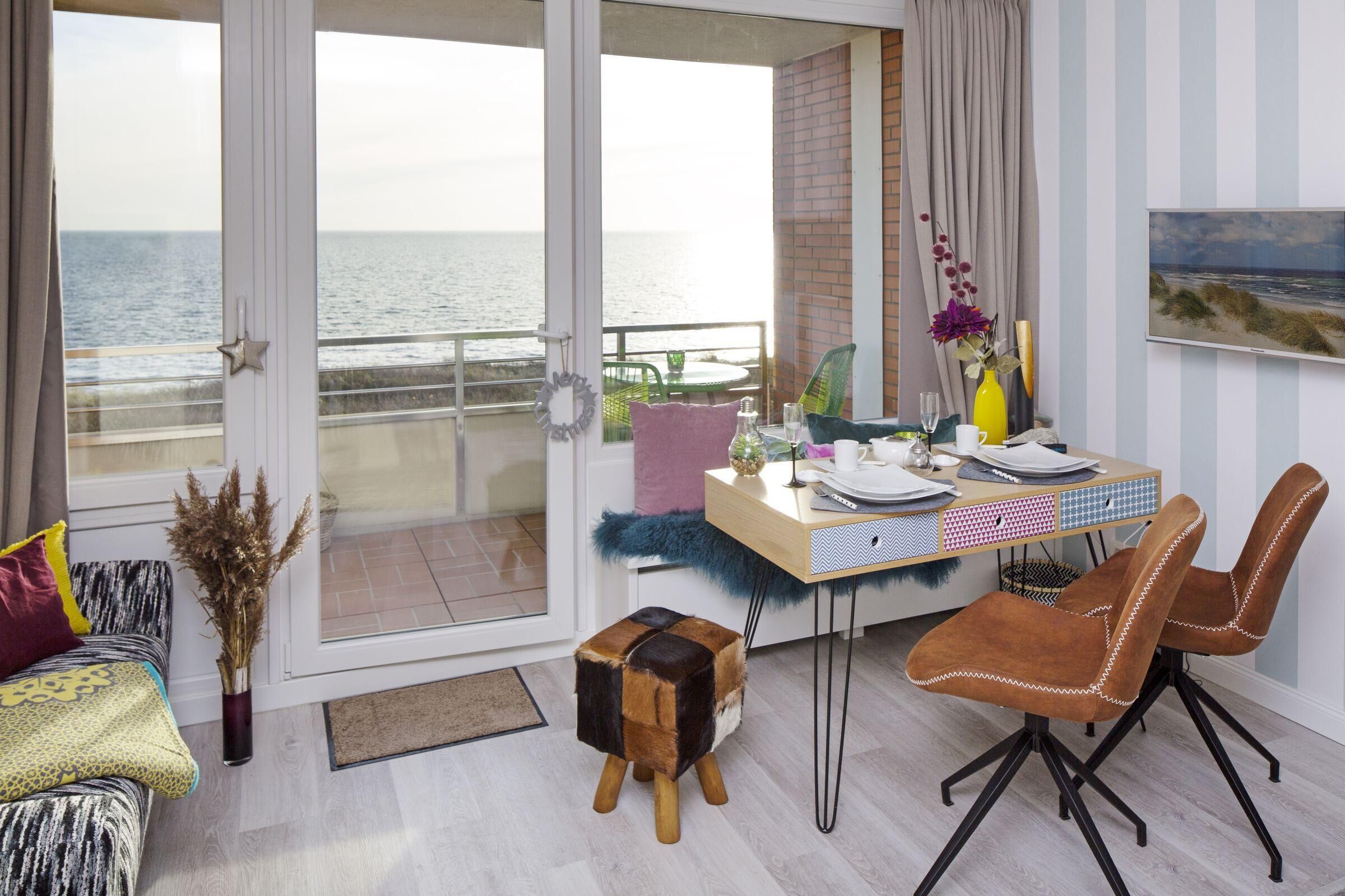 Ein modernes Wohnzimmer mit Balkon und Meerblick, einer Essecke mit gestreifter Tapete und einer gemütlichen Sitzecke mit einem gemusterten Sofa.