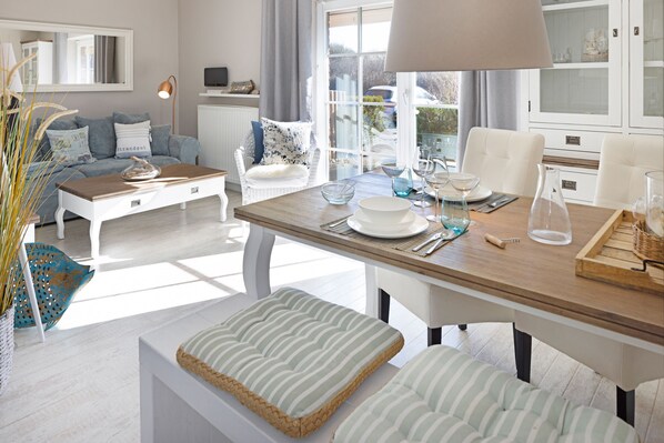 A modern living room with a wooden dining table, white sofas, and a large window with a view of the outdoors.