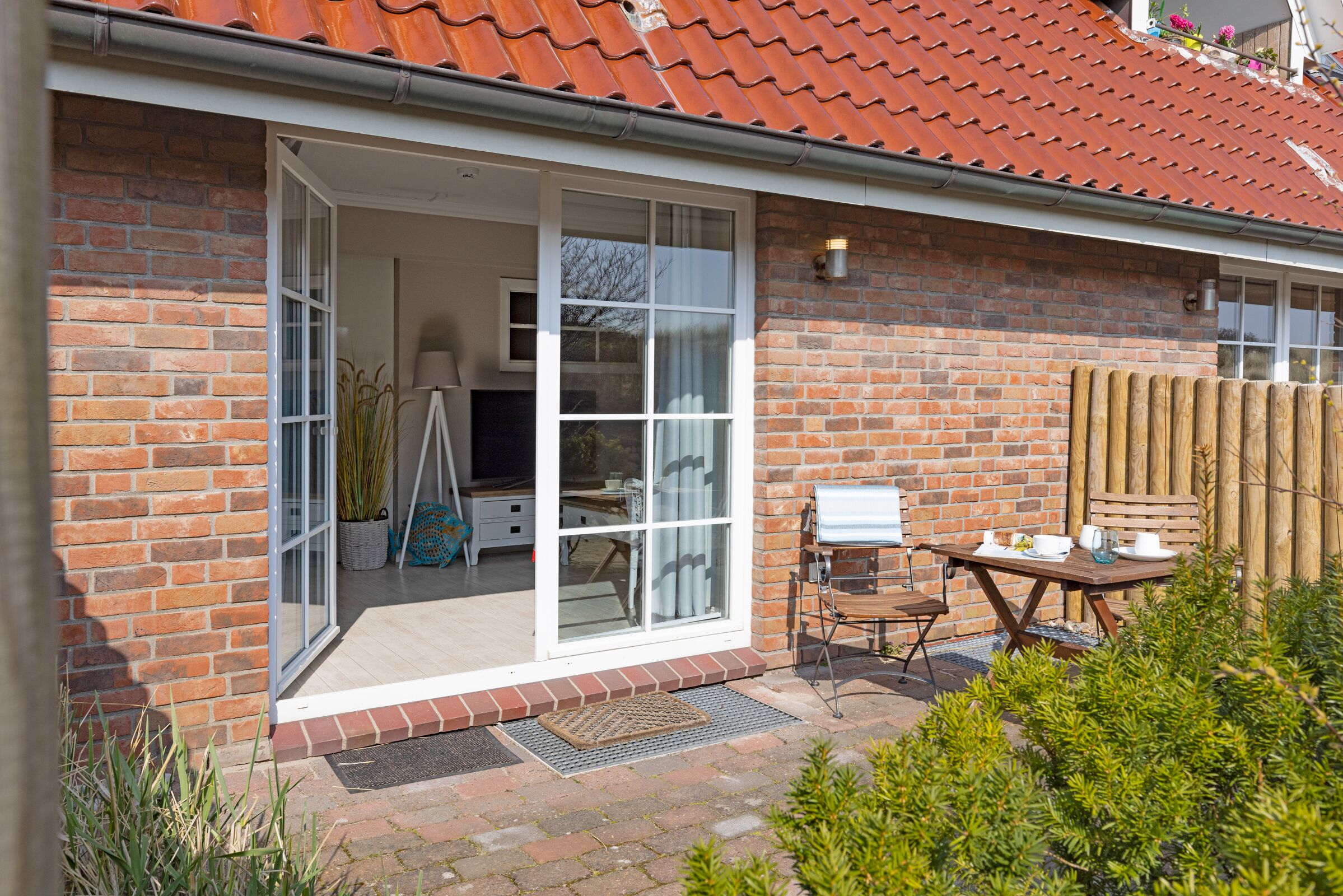Ein Ziegelhaus mit rotem Ziegeldach, eine Glastür, die zu einem Raum mit einem Bett und einem Schreibtisch führt, und eine kleine Terrasse mit Tisch und Stühlen.