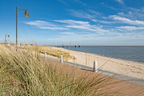 Ein Küstenweg mit einer Lampe und Gras im Vordergrund, der zu einem Sandstrand mit dem Meer im Hintergrund führt.