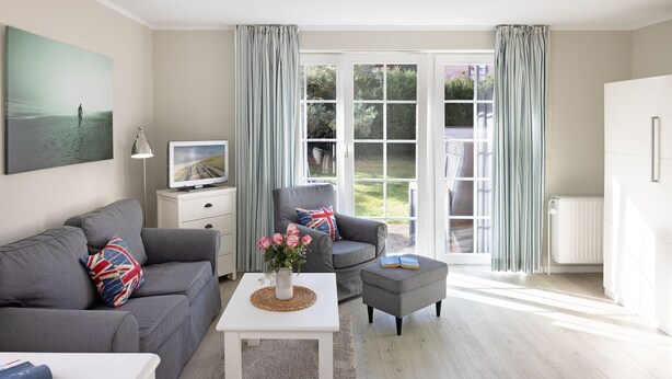 A modern living room with a grey sofa, a white coffee table, and a grey armchair. There is a television on a white stand, a small white table with a vase of flowers, and a book on the armchair. The room has a large window with curtains, a white radiator, and a door leading outside.