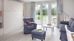 Ein modernes Wohnzimmer mit einem grauen Sofa, einem passenden Hocker und einem Sessel mit einem Kissen mit Wimpelmotiv. Ein kleiner Tisch mit einem Buch und einer Lampe steht in der Mitte.