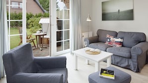 Ein modernes Wohnzimmer mit einem grauen Sofa, einem weißen Couchtisch und einem grauen Sessel. Eine Glastür führt auf eine Terrasse mit Tisch und Stühlen.