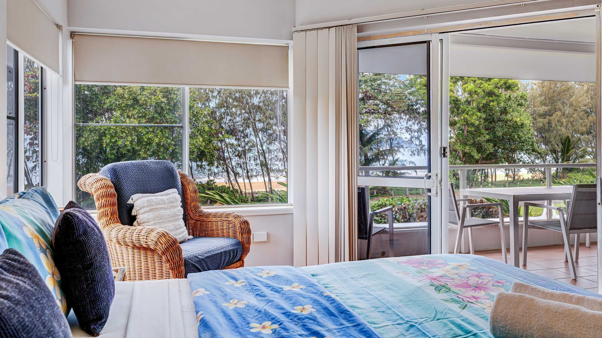 A room with a bed, a wicker chair, a table, and a sliding glass door leading to an outdoor area.