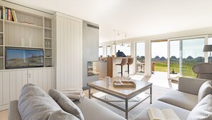 A modern living room with a large flat-screen TV, a wooden coffee table, and a sofa. There is a bookshelf with books and decorative items. The room has large windows with a view of the outdoors.