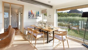 A dining area with a wooden table set for six, surrounded by six wooden chairs with cushions. There is a large window providing a view of a garden and a building outside.