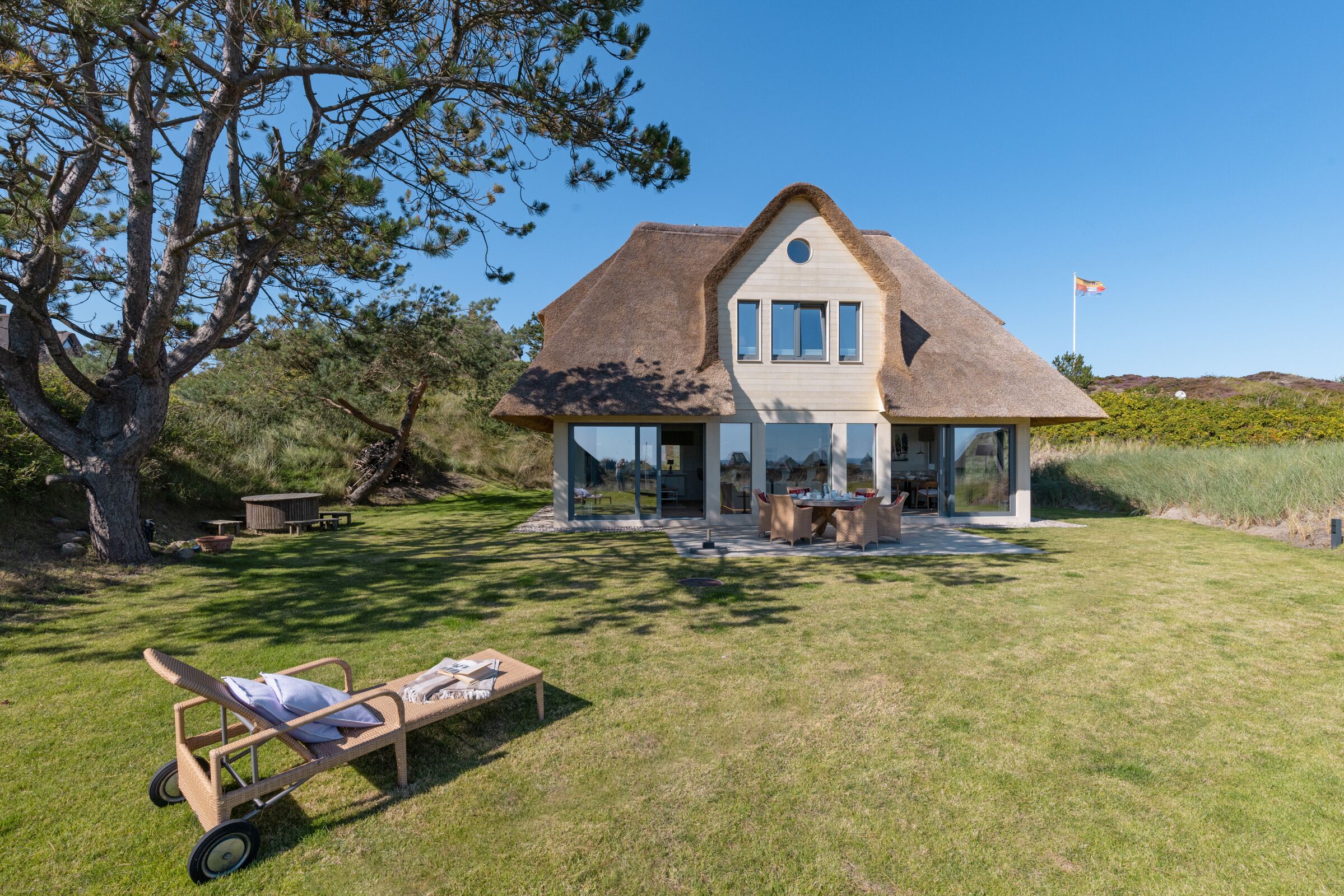 A house with a thatched roof, a large glass window, and a lawn with a sun lounger.