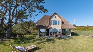 A house with a thatched roof, a large glass window, and a lawn with a sun lounger.