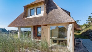 A thatched-roof house with a glass door and windows, surrounded by grass and a stone pathway.