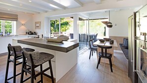 A modern kitchen with a central island, bar stools, a dining area with a table and chairs, and a large window offering a view of the outdoors.