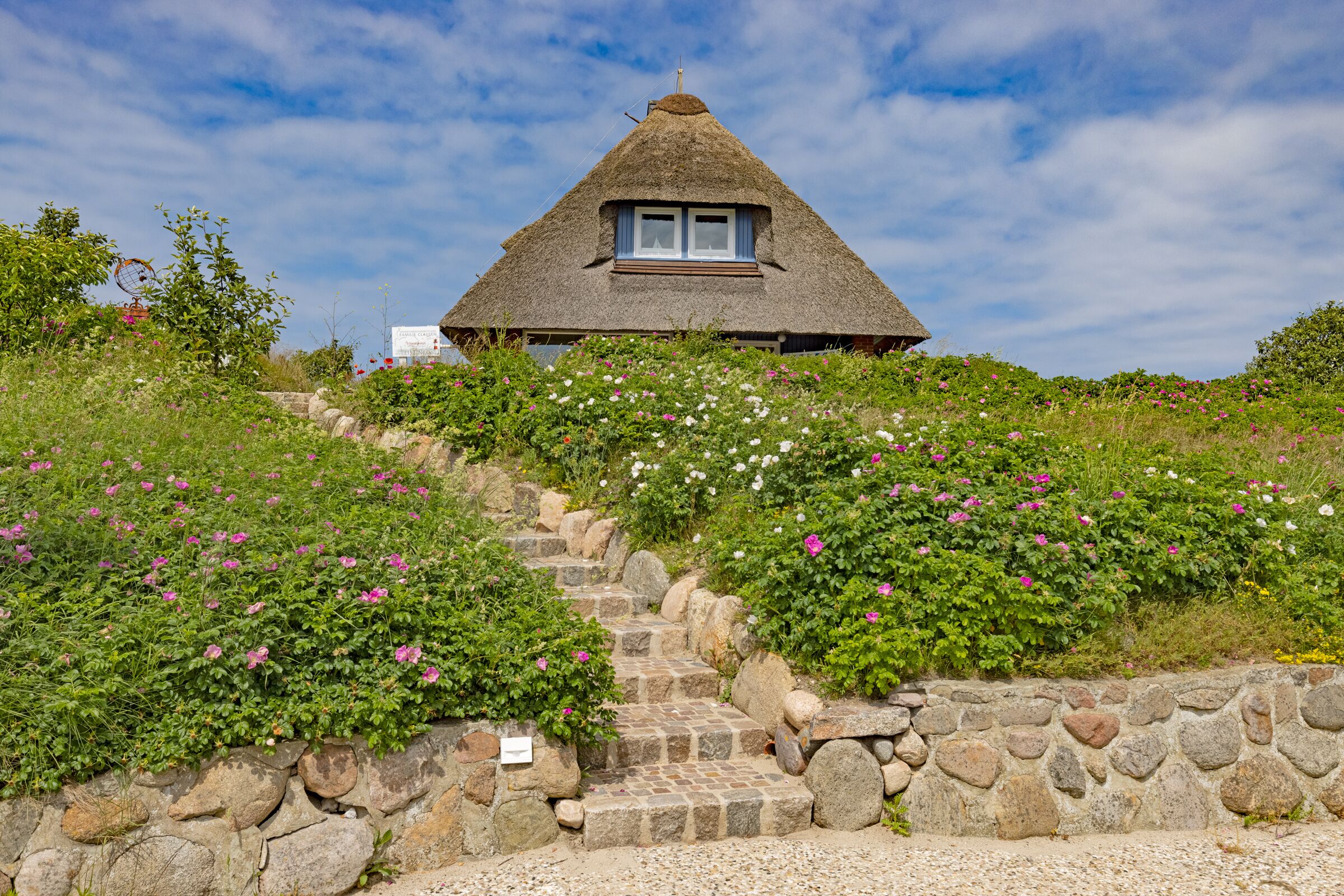 Una casa con techo de paja y una escalera de piedra que lleva a ella, rodeada de abundante vegetación y flores.