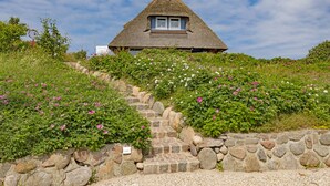 Ein Haus mit einem Strohdach und einer Steintreppe, die zu ihm hinaufführt, umgeben von üppigem Grün und Blumen.